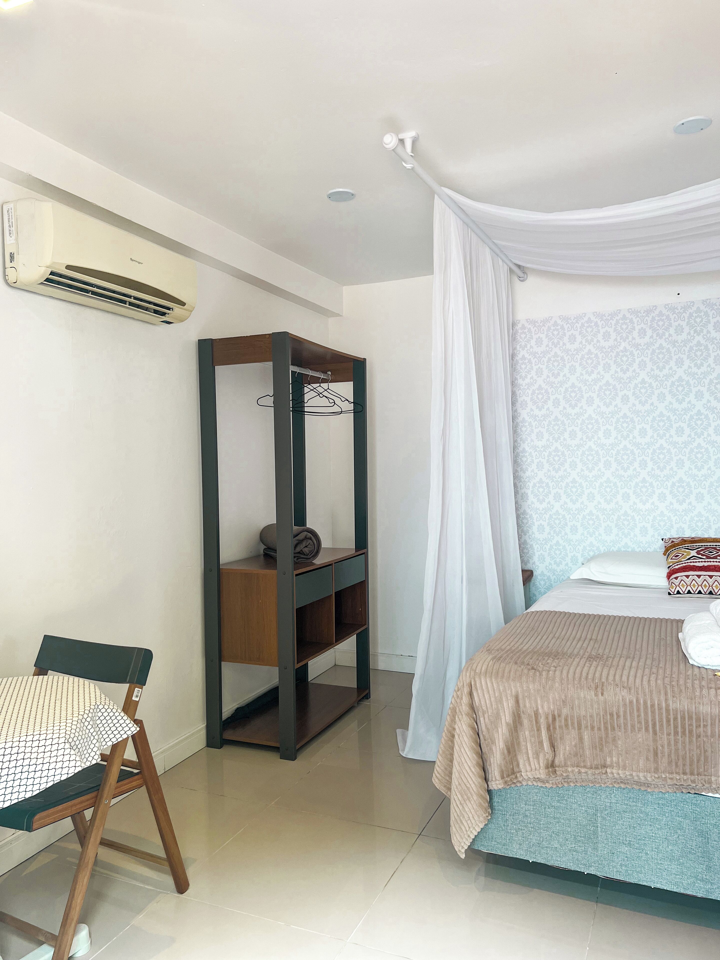 A bedroom with a bed, a wooden chair, a small wooden shelf, a white curtain, and an air conditioning unit on the wall.