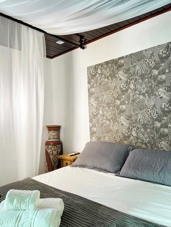 Un dormitorio con cama, almohadillas grises, una pared con diseño, una mesita de noche de madera, un jarrón y una cortina.