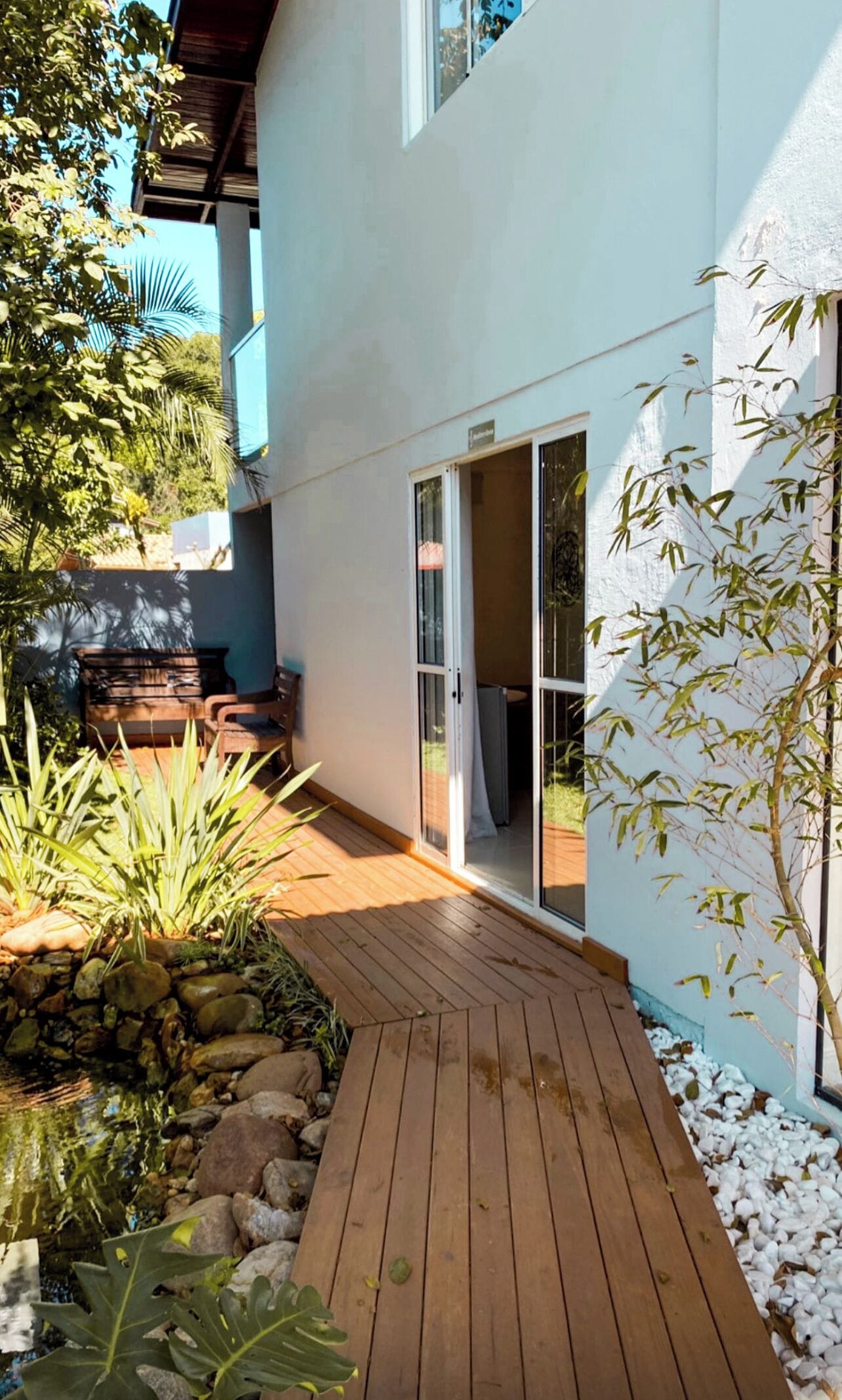 A wooden deck leading to a sliding glass door, surrounded by greenery and a stone wall.