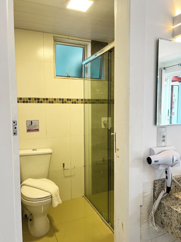 A bathroom with a toilet, a glass shower enclosure, and a hairdryer mounted on the wall. - Essência do Sol Pousada (Florianópolis)