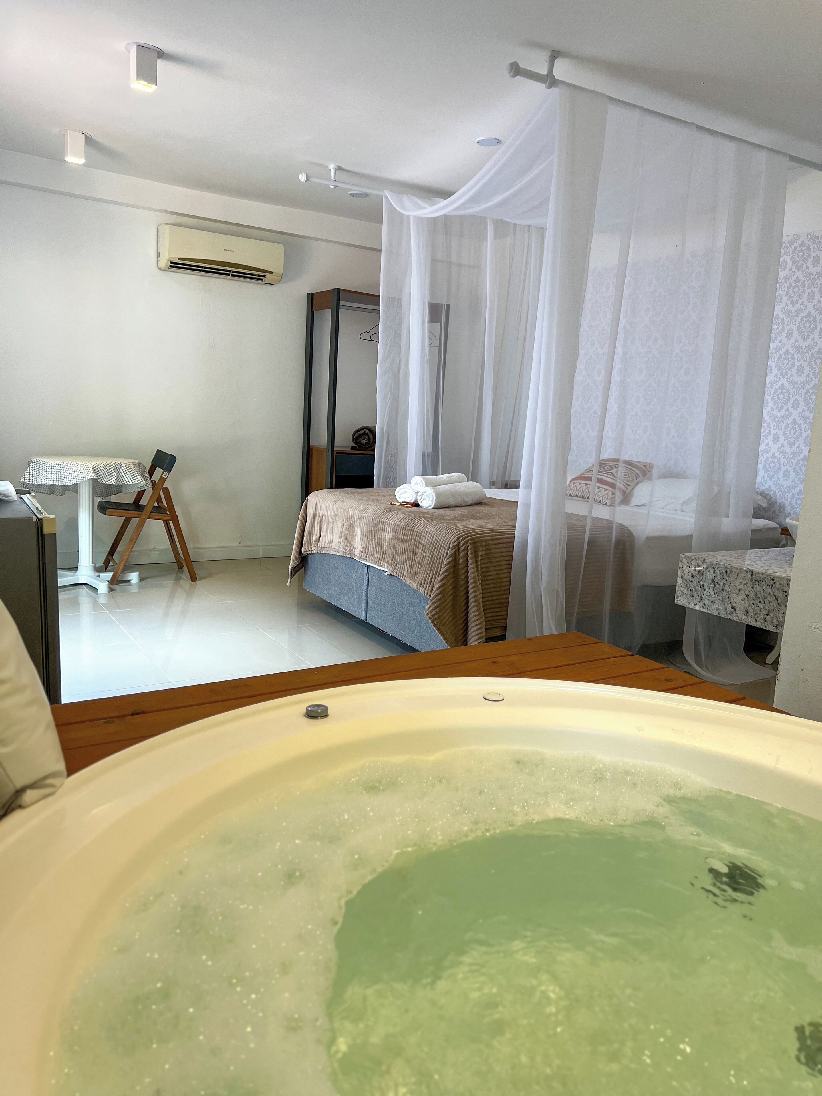 A hotel room with a large bathtub, a bed with a canopy, a desk with a chair, and an air conditioning unit.