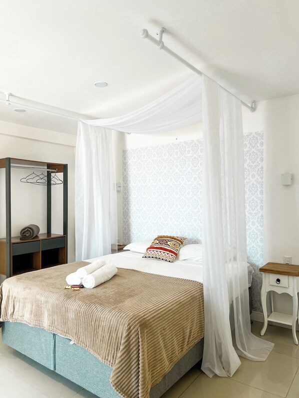 A neatly made bed with a beige blanket, a decorative pillow, and white towels. There is a wooden nightstand with a drawer, a mirror, and a hanging coat rack. A white canopy is draped over the bed. - Essência do Sol Pousada (Florianópolis)
