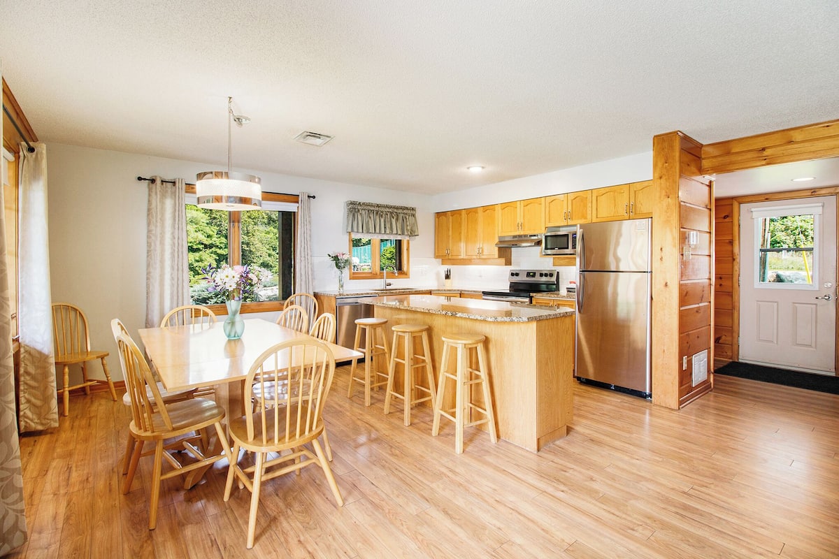 Una cocina moderna con armarios de madera, una isla central con taburetes, un refrigerador de acero inoxidable y un comedor con sillas y una mesa de madera.