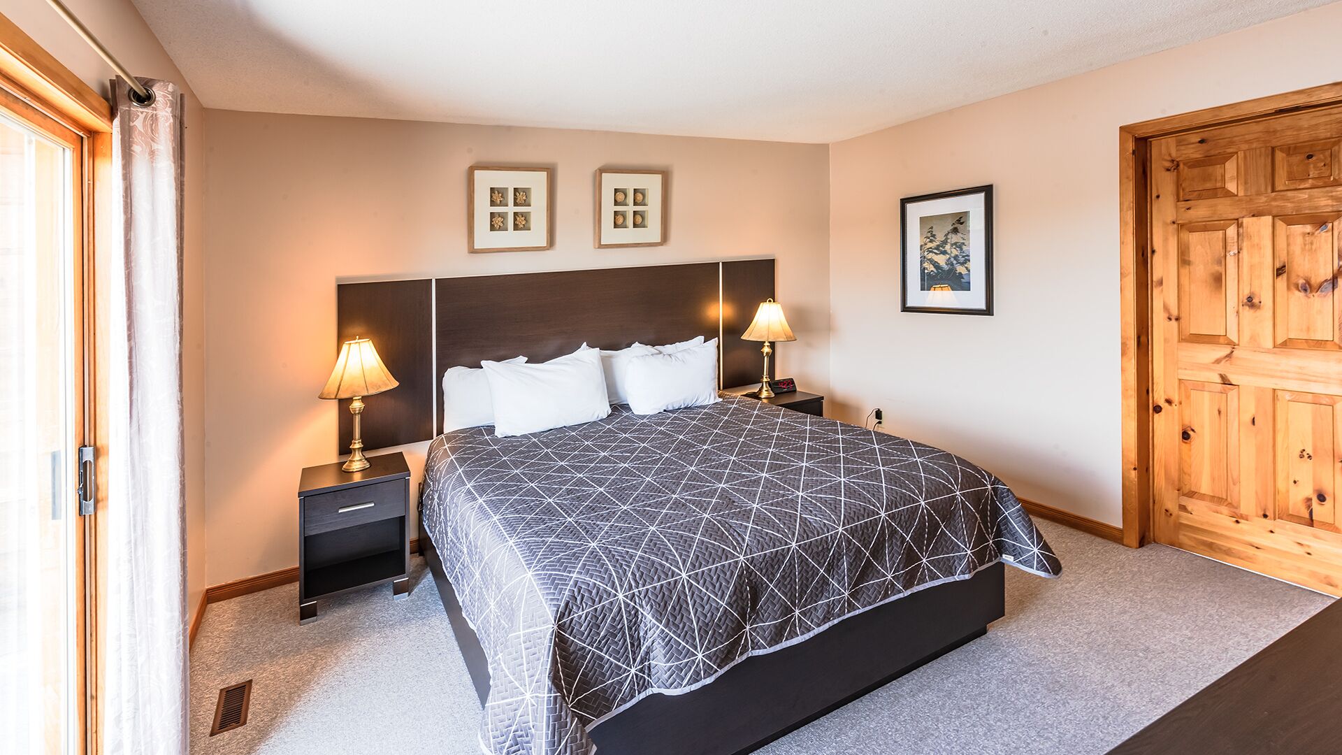 A bedroom with a large bed, two bedside tables with lamps, a wooden door, and framed pictures on the wall.