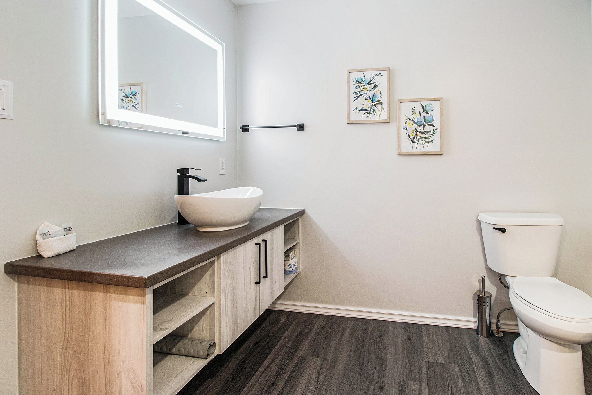 Une salle de bain avec un lavabo blanc, un meuble-lavabo en bois foncé et des toilettes blanches. Deux œuvres d’art florales encadrées ornent le mur.