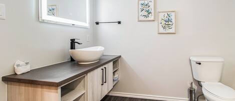 Une salle de bain avec un lavabo blanc, un meuble-lavabo en bois foncé et des toilettes blanches. Deux œuvres d’art florales encadrées ornent le mur.