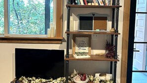 A bookshelf with books and decorative items, a fireplace with a framed picture, and a window with a view of trees.