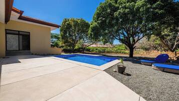 Una piscina con un borde de baldosas, un pequeño árbol y una silla de descanso azul.