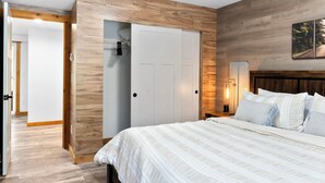 A bedroom with a bed, wooden wall paneling, a white door, and a nightstand with a lamp.