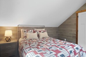 Une chambre avec un plafond en pente, une tête de lit en bois, un lit recouvert d’une courtepointe à motifs, une table de chevet avec une lampe et une commode de chevet avec des tiroirs.