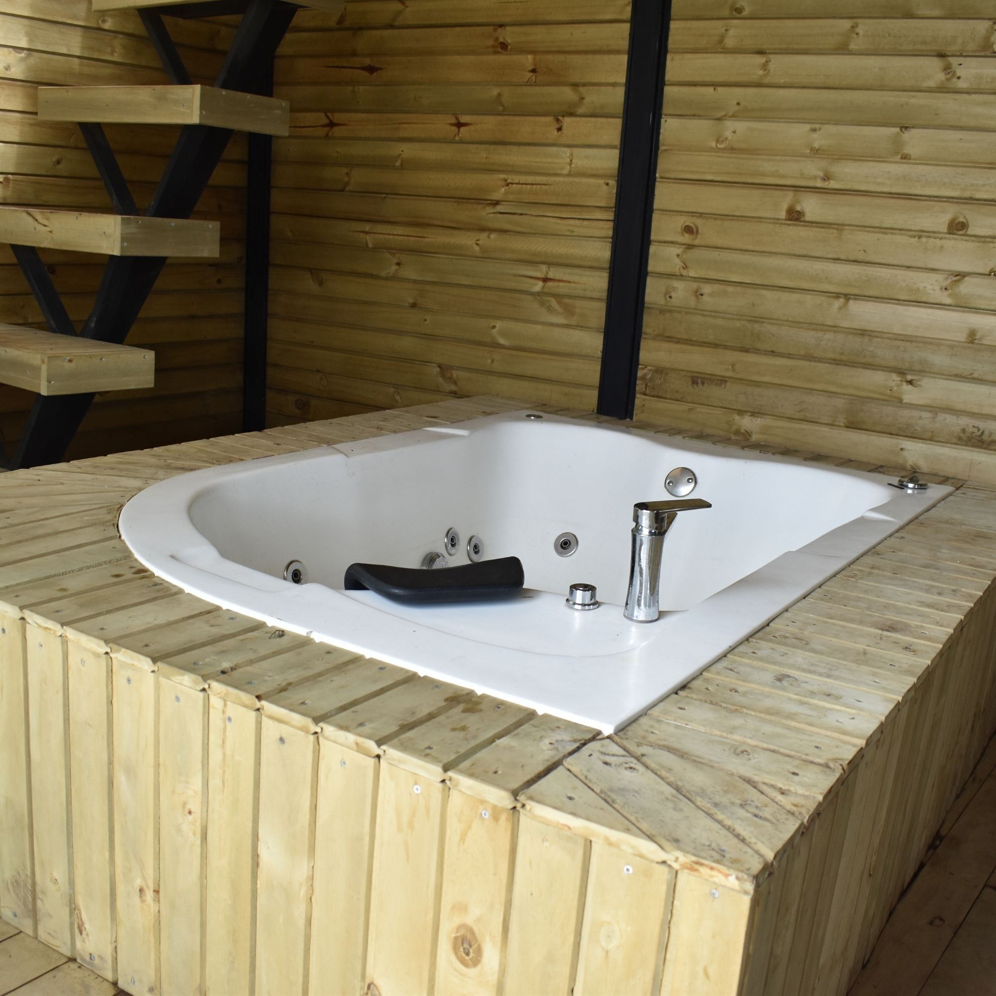 A wooden spa tub with a black seat and a chrome faucet, set against a wooden wall with a ladder.