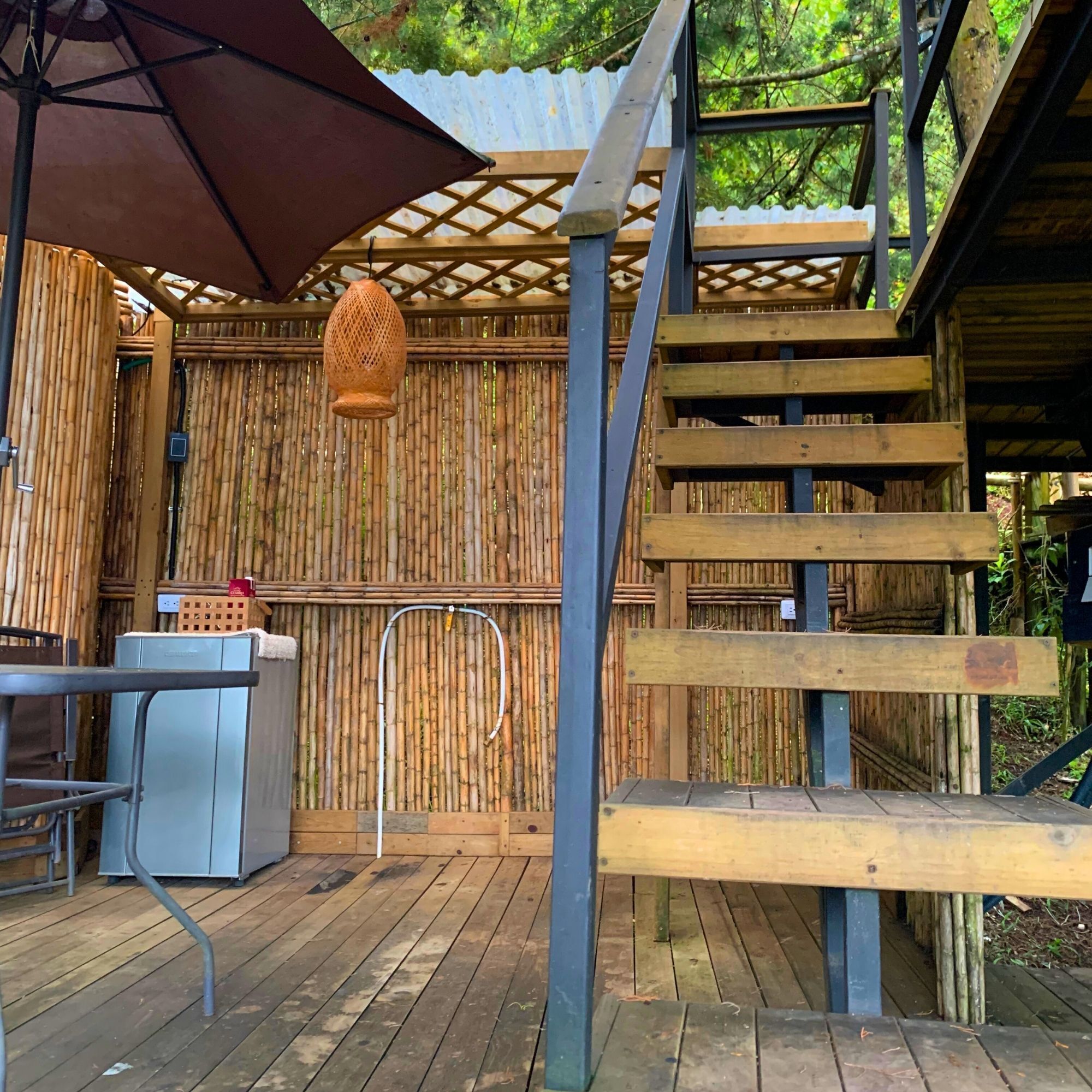 Una terraza de madera con una mesa y sillas de metal, una lámpara colgante y un muro de bambú.