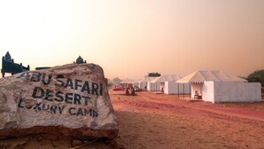 A desert landscape with a large rock inscribed with "ABU SAFARI DESERT LUXURY CAMP" and several white tents in the background. - Abu Safari   (Jaisalmer)