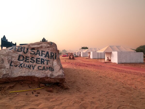 Paisaje desértico con una gran roca que muestra la inscripción "ABU SAFARI DESERT LUXURY CAMP" y varias tiendas blancas al fondo.