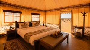 A room with a large bed, a bench, a nightstand, a lamp, and a view of the desert outside. - Abu Safari   (Jaisalmer)