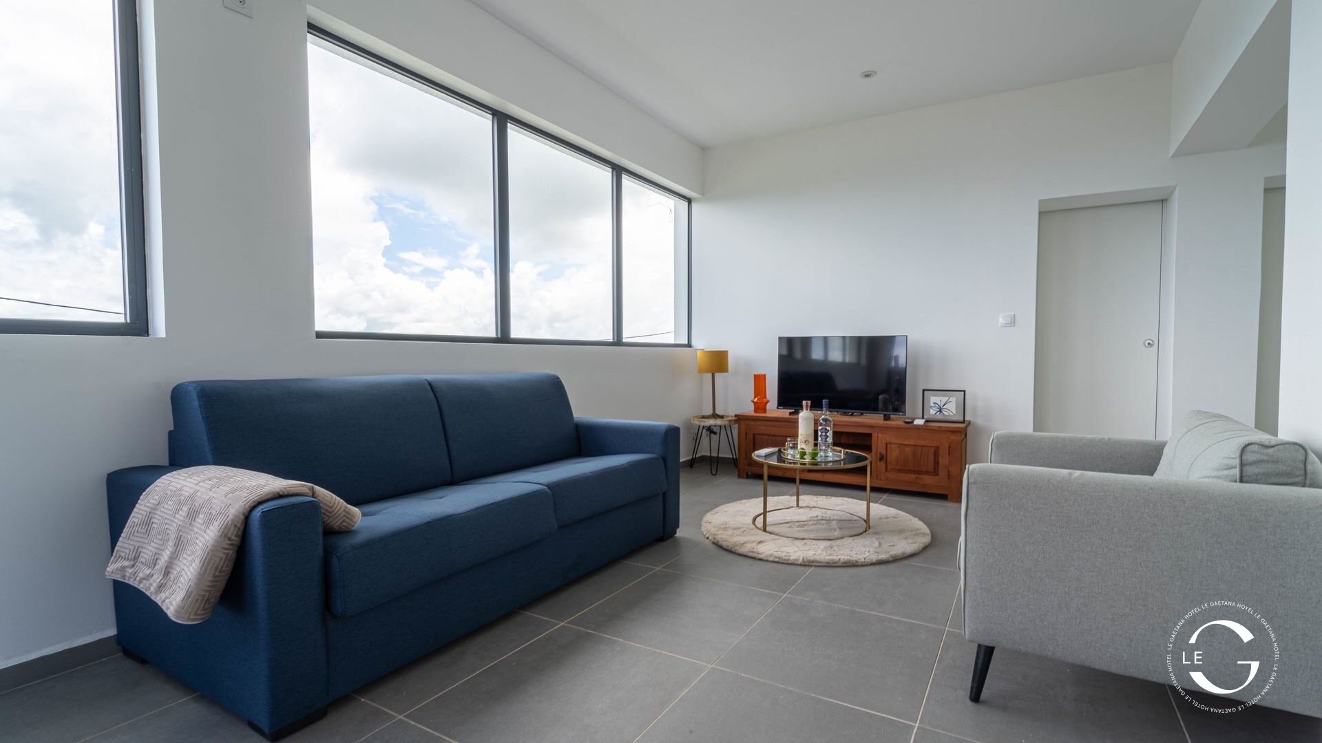 Una sala de estar moderna con un sofá azul, un sillón gris, un televisor y un ventanal con vistas al cielo.