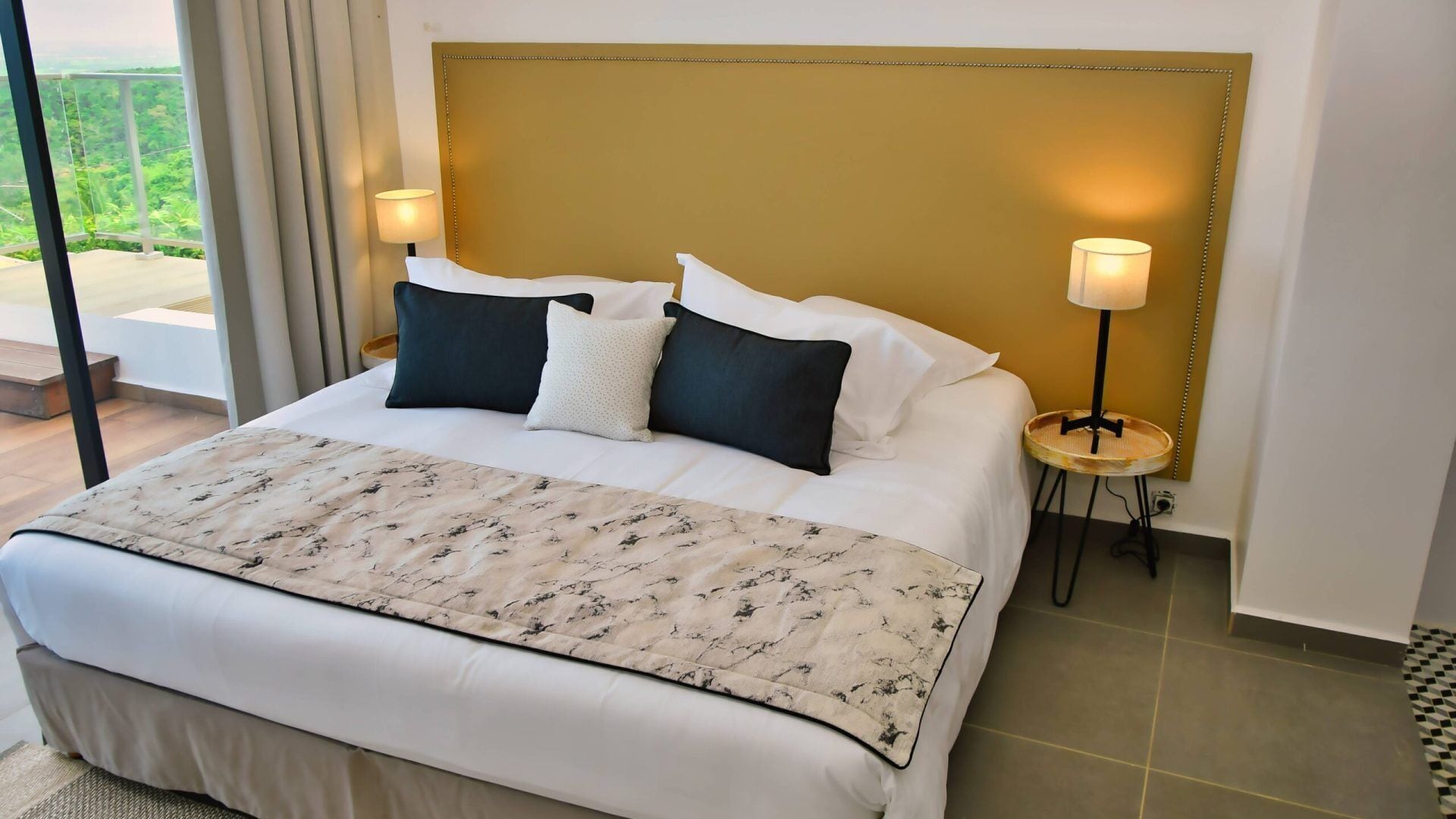 Un grand lit avec une tête de lit dorée, une literie blanche et un couvre-lit à motifs. Il y a deux lampes de chevet et une petite table ronde avec une lampe.