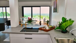 A modern kitchen with a large island, a view of greenery and a bridge, and a dining area with chairs and a table set for two. - Le Gaetana hôtel (Sainte-Rose)