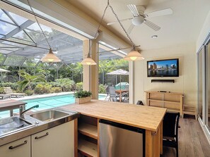 A modern kitchen with a large island, a built-in sink, and a stainless steel refrigerator. There is a ceiling fan, pendant lights, and a large window overlooking a pool area. - Heated Pool Home Near Bonita Beach w/ Arcade Room (Bonita Springs)