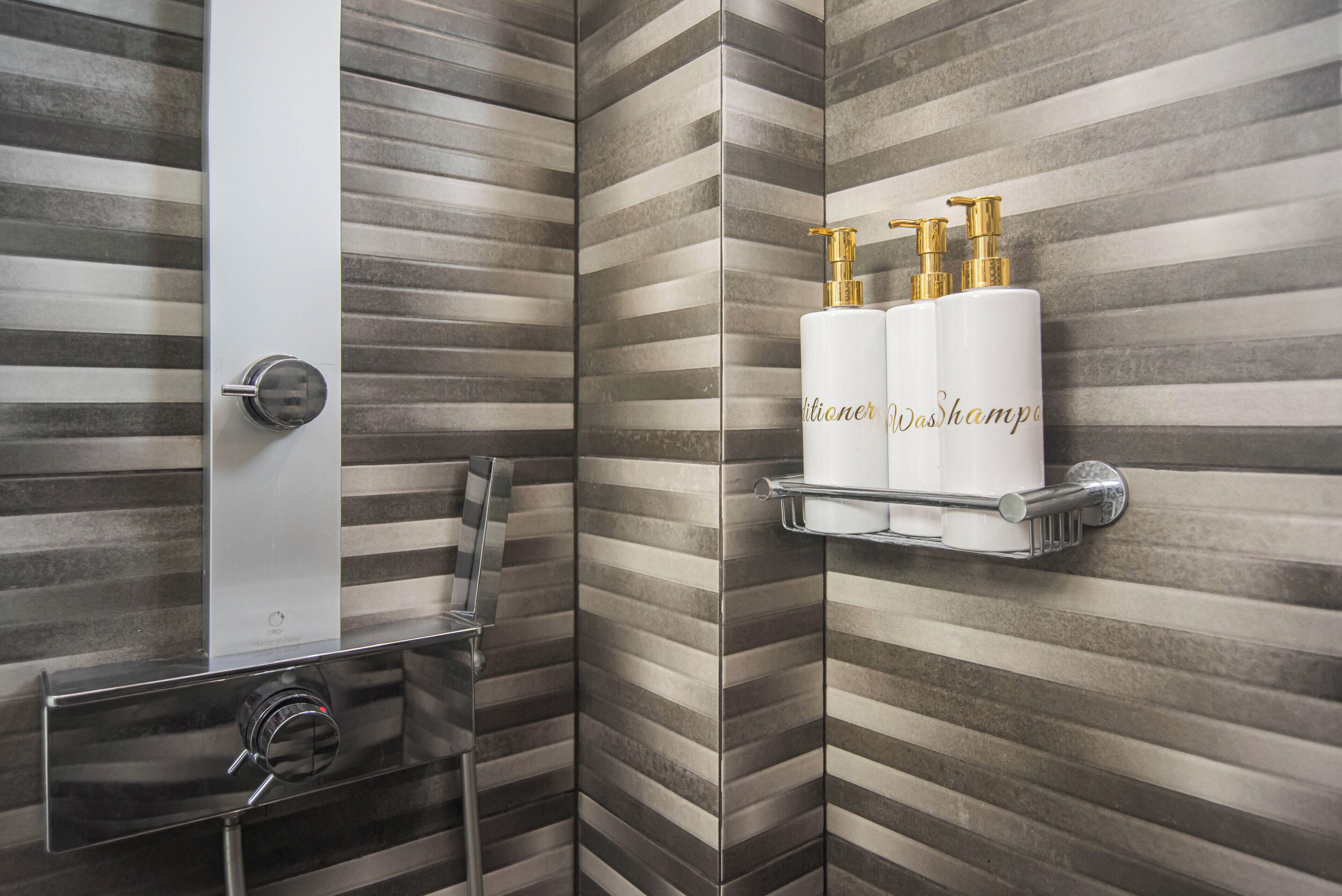 Un baño moderno con paredes de azulejos a rayas, recinto de ducha cromado y repisa con dos botellas de loción.