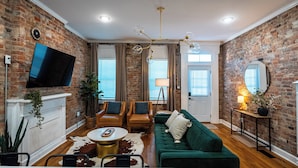 A living room with a brick wall, a green sofa, two armchairs, a coffee table, and a TV mounted on the wall.
