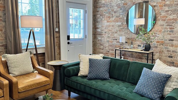 A cozy living room with a green sofa, leather armchair, and a round coffee table. A brick wall and a large mirror are visible.