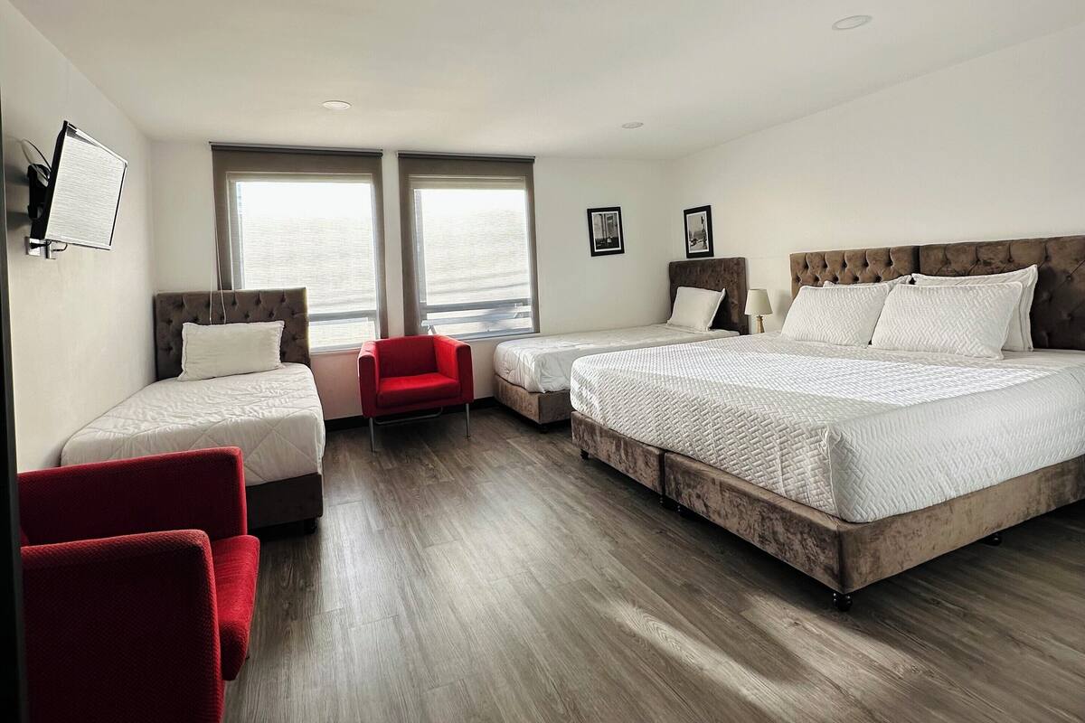Una habitación de hotel moderna con dos camas individuales, un sillón rojo y una cama grande con cabecero de madera.