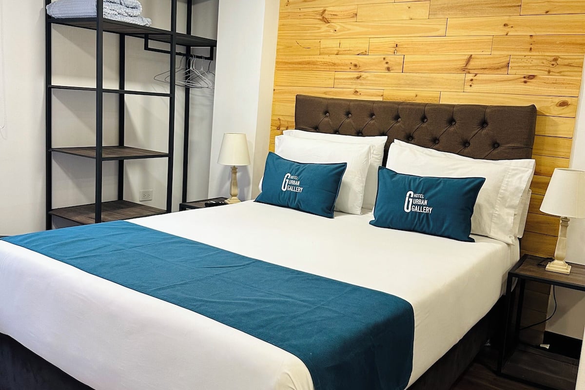 Habitación de hotel con cama, cabecera de madera, mesita de noche, lámpara, estante con ropa doblada y estante en la pared.