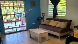 Un salon comprenant un canapé, une table basse, un ventilateur de plafond et une vue sur l’extérieur à travers une fenêtre avec des rideaux.