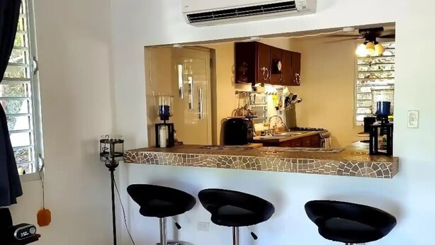 Une cuisine avec un mur recouvert de carreaux, trois tabourets de bar noirs et un appareil de climatisation placé au-dessus du comptoir.