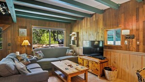 A cozy living room with a sofa, a coffee table, a TV, and a window with a view of greenery. - Rainier's Iconic Cabin with Forest Views-WiFi-Hot Tub (Ashford)