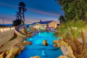 Une piscine avec un toboggan, entourée de rochers et de palmiers, avec une maison en arrière-plan.