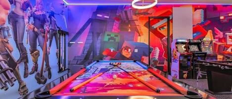 Una mesa de billar con un diseño abstracto y colorido, rodeada de máquinas de videojuegos y una pared con ilustraciones inspiradas en Minecraft.