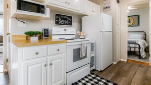 Une cuisine compacte avec des meubles blancs, un four à micro-ondes, une cuisinière et un réfrigérateur. Un panneau est accroché au mur avec l’inscription : « DONNEZ-NOUS AUJOURD’HUI NOTRE PAIN DE CHAQUE JOUR. » Une petite plante et un panneau d’accueil sont posés sur le plan de travail.