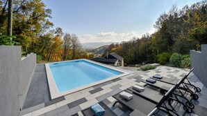 En rektangulär pool med ett rutmönstrat poolområde, solstolar och utsikt över ett bergigt landskap.