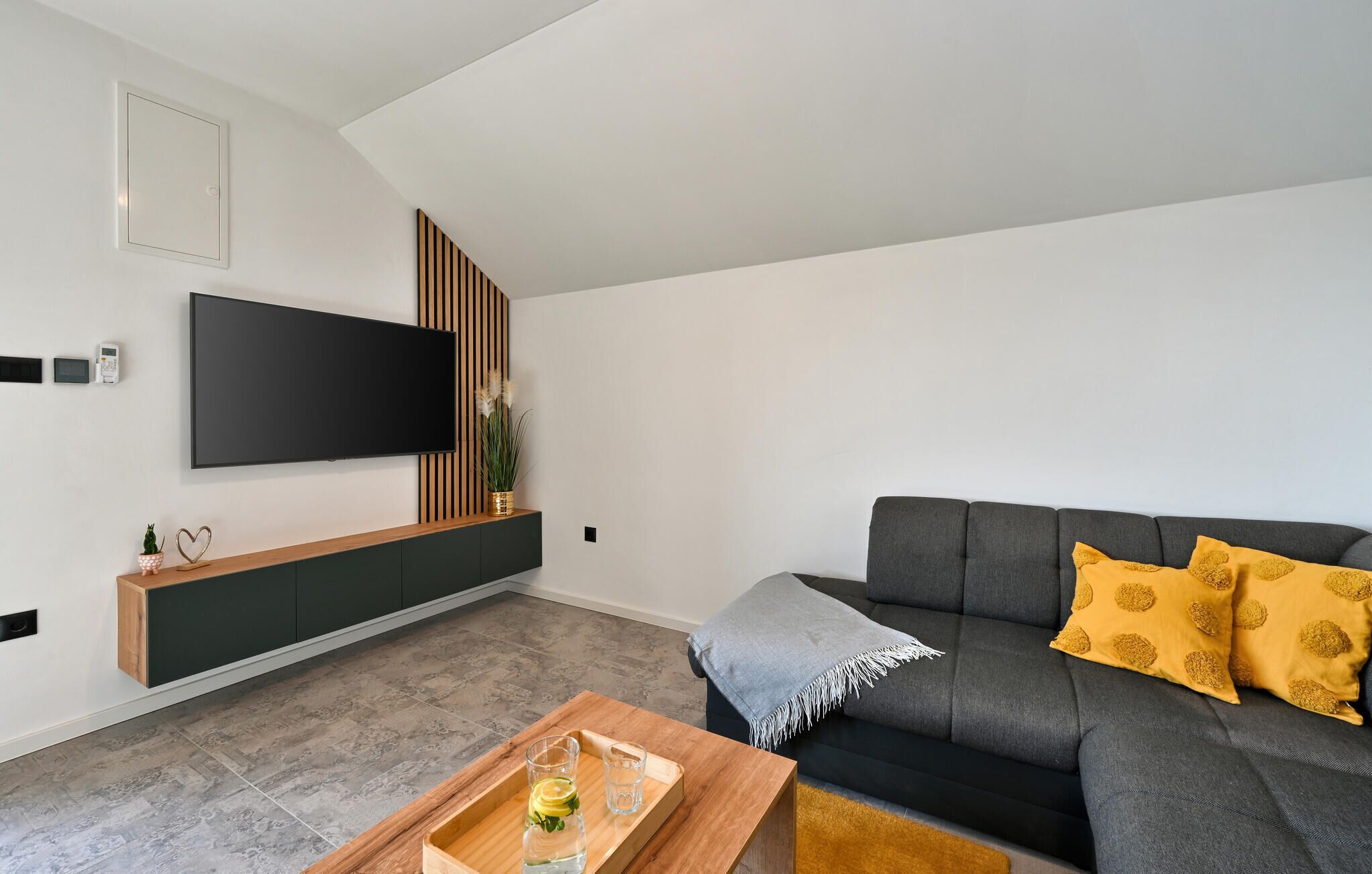 Ein modernes Wohnzimmer mit einem an der Wand befestigten Flachbildfernseher, einem grauen Sofa mit gelben Kissen, einem Holztisch mit Gläsern und einer Serviertablett sowie einer kleinen Pflanze auf einer Holzkonsole.