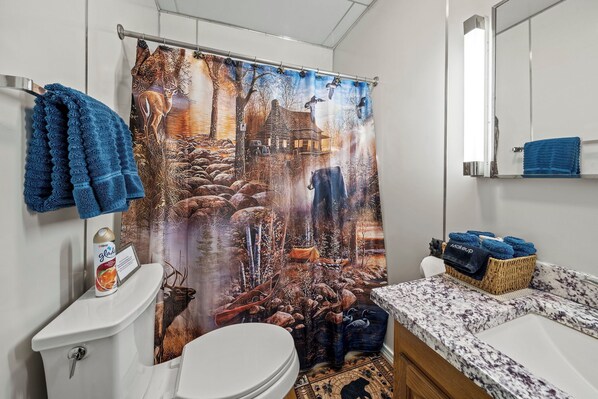 A bathroom with a deer and forest scene shower curtain, a toilet, a sink with a marble countertop, and a basket of towels.