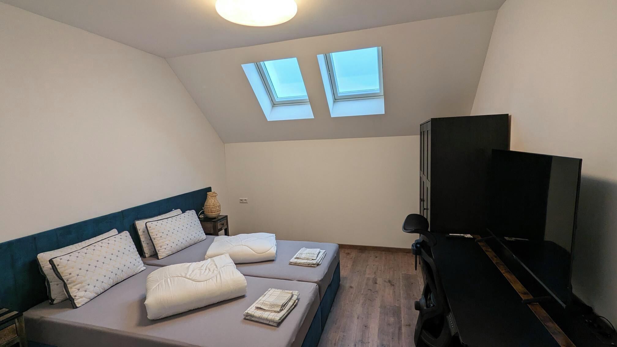 A bedroom with a bed, two pillows, a bedside table with a lamp, a TV, and two skylights.