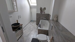 A modern bathroom with a bathtub, toilet, and sink. There is a skylight above the toilet and a small plant on the wall.