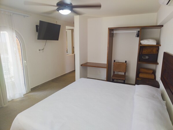 A hotel room with a bed, a ceiling fan, a TV mounted on the wall, and a small seating area with a chair and shelves.