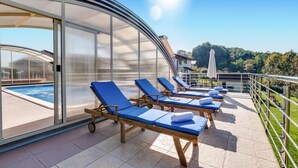 Un espacio al aire libre iluminado por el sol, con piscina, sillones reclinables azules y una estructura de vidrio.
