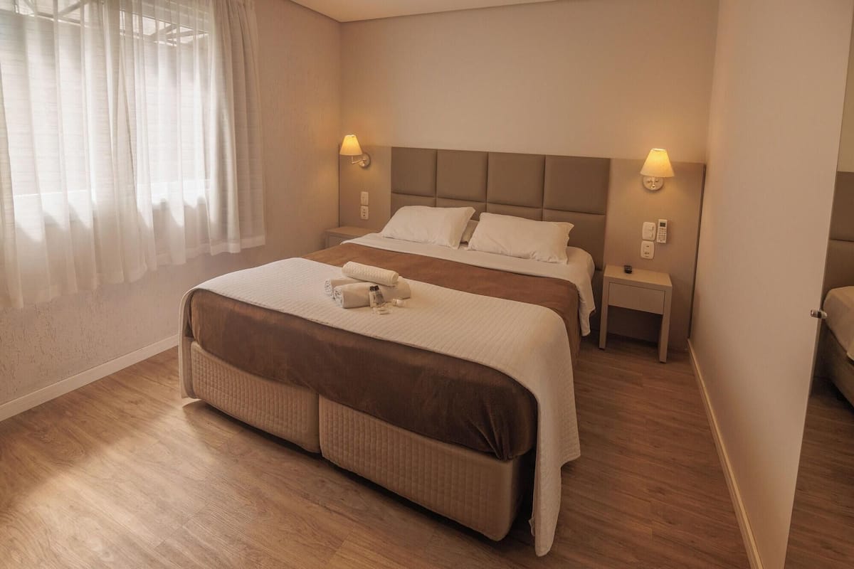 A double bed with a brown headboard, two bedside tables with lamps, and a window with sheer curtains.