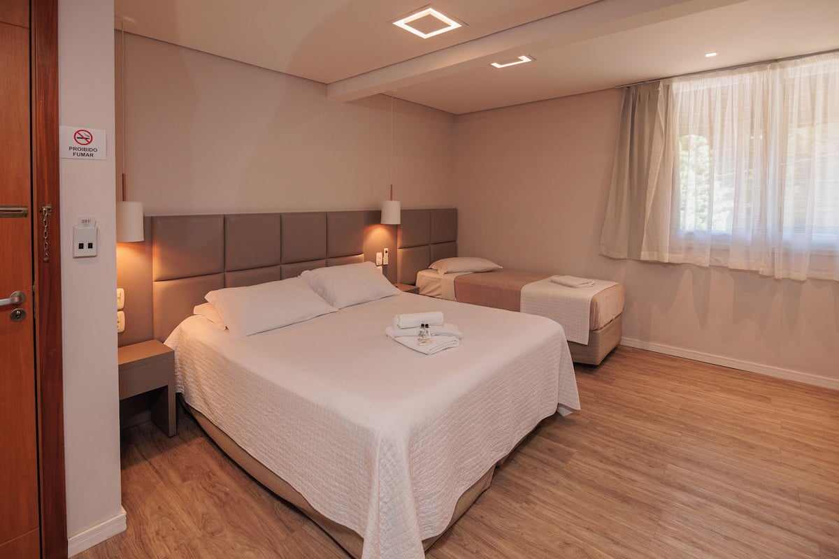 A hotel room with a large bed, bedside tables, a nightstand, a window with sheer curtains, and a door with a "No Smoking" sign.