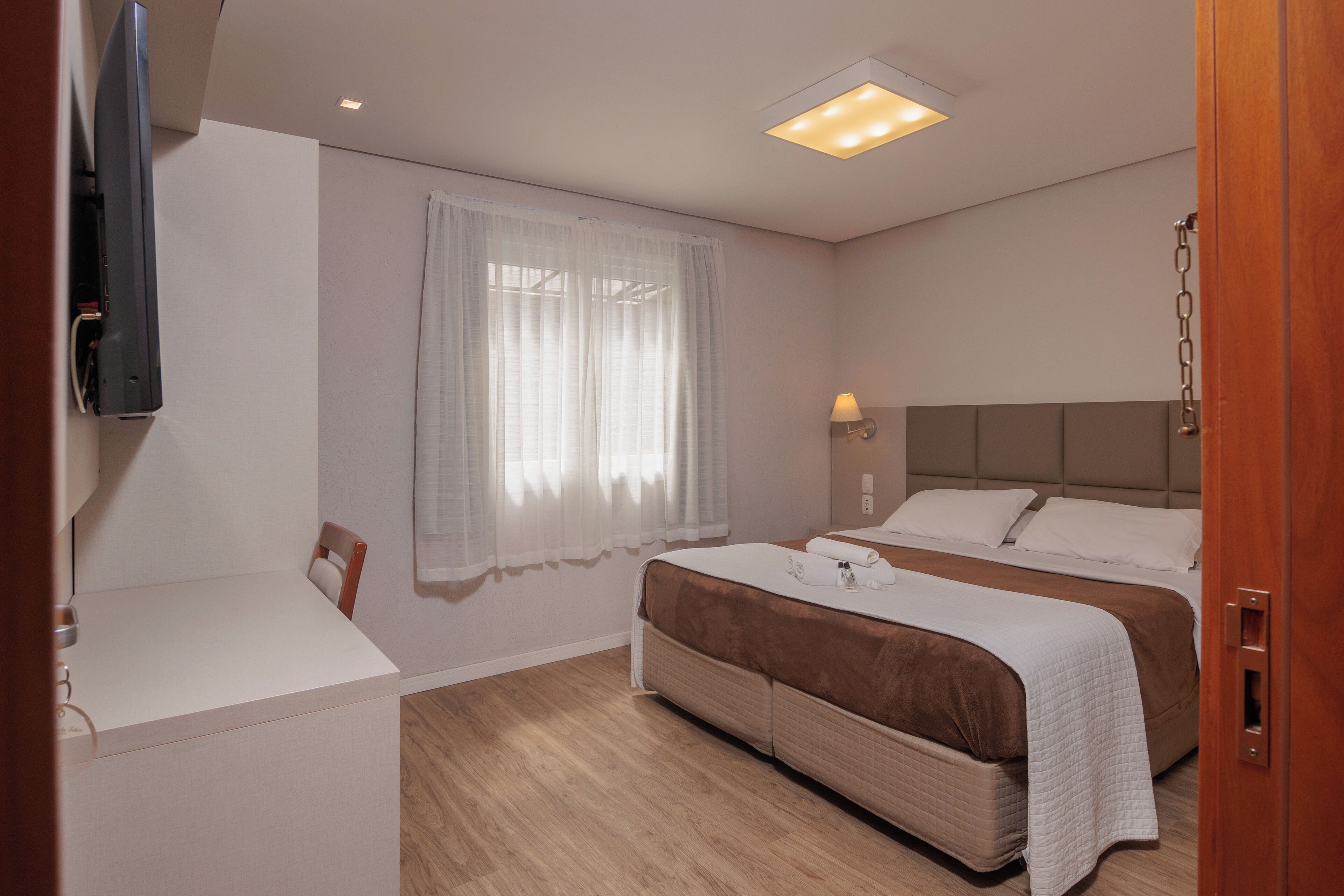 Camera d'albergo con due letti, una TV a parete, una lampada da comodino e una finestra con tende leggere.