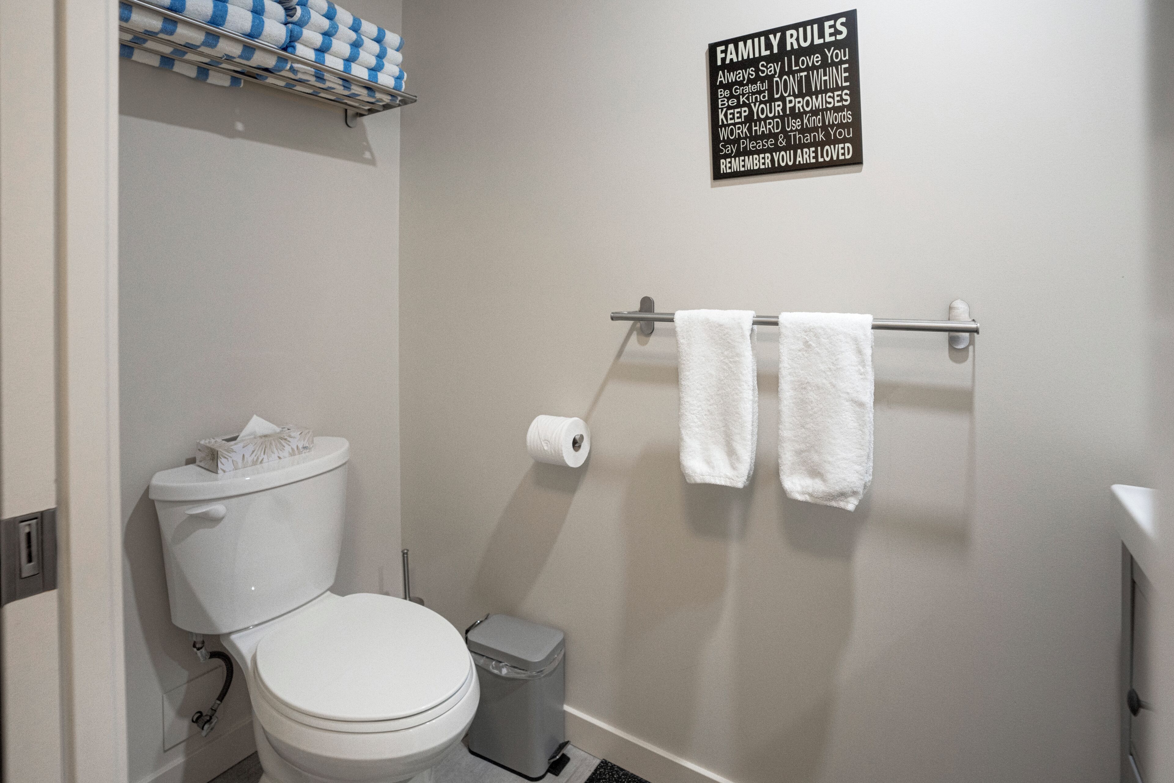 A bathroom with a toilet, a trash bin, a towel rack with two towels, and a sign with family rules.