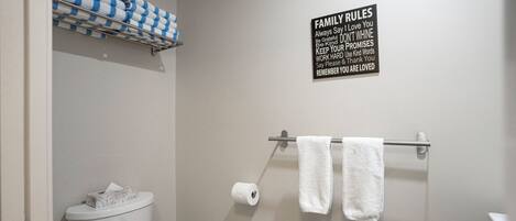 A bathroom with a toilet, a trash bin, a towel rack with two towels, and a sign with family rules.