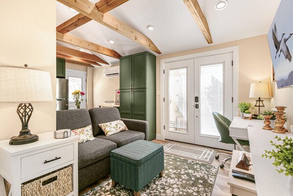 A cozy living area with a sofa, ottoman, and a desk with a chair. There is a door leading to a balcony, and a lamp on a side table.