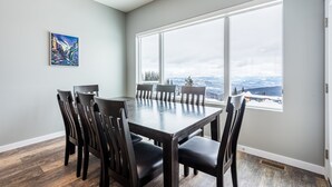 A dining area with a large table and chairs, a painting on the wall, and a view of a snowy landscape through large windows.