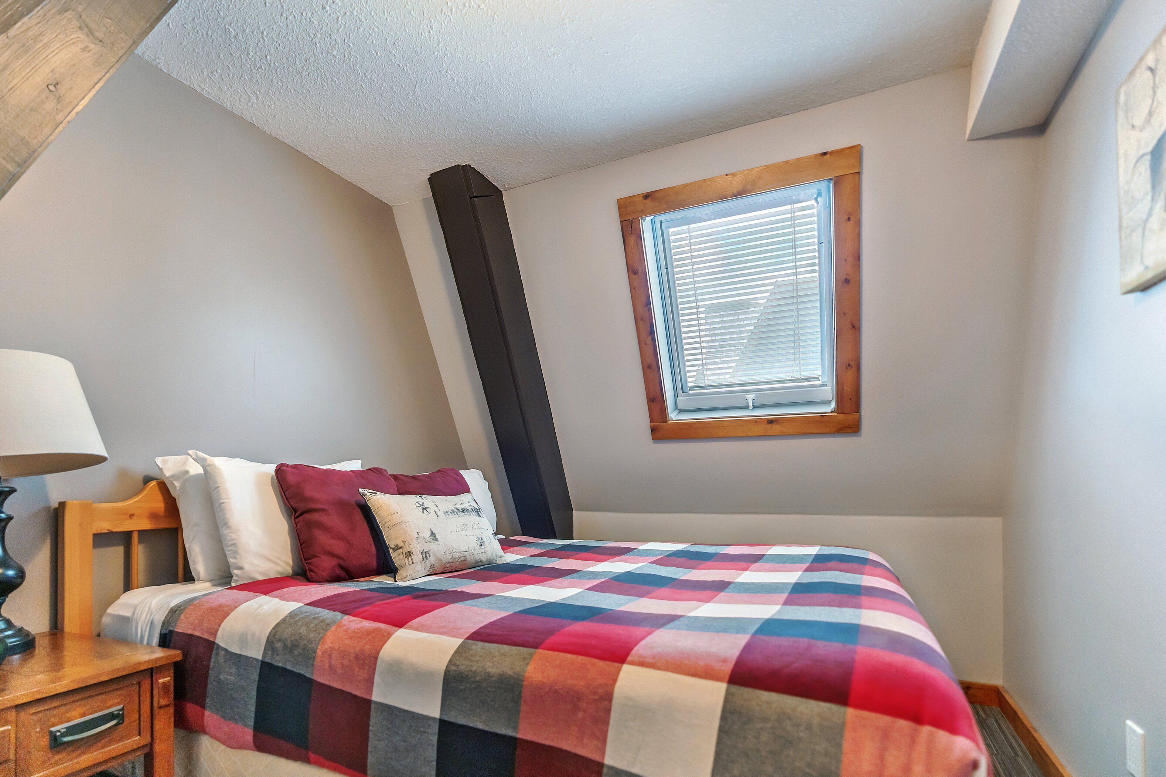 Un dormitorio con una colcha a cuadros, una mesita de noche con lámpara, una ventana con persianas y un cabecero de madera.