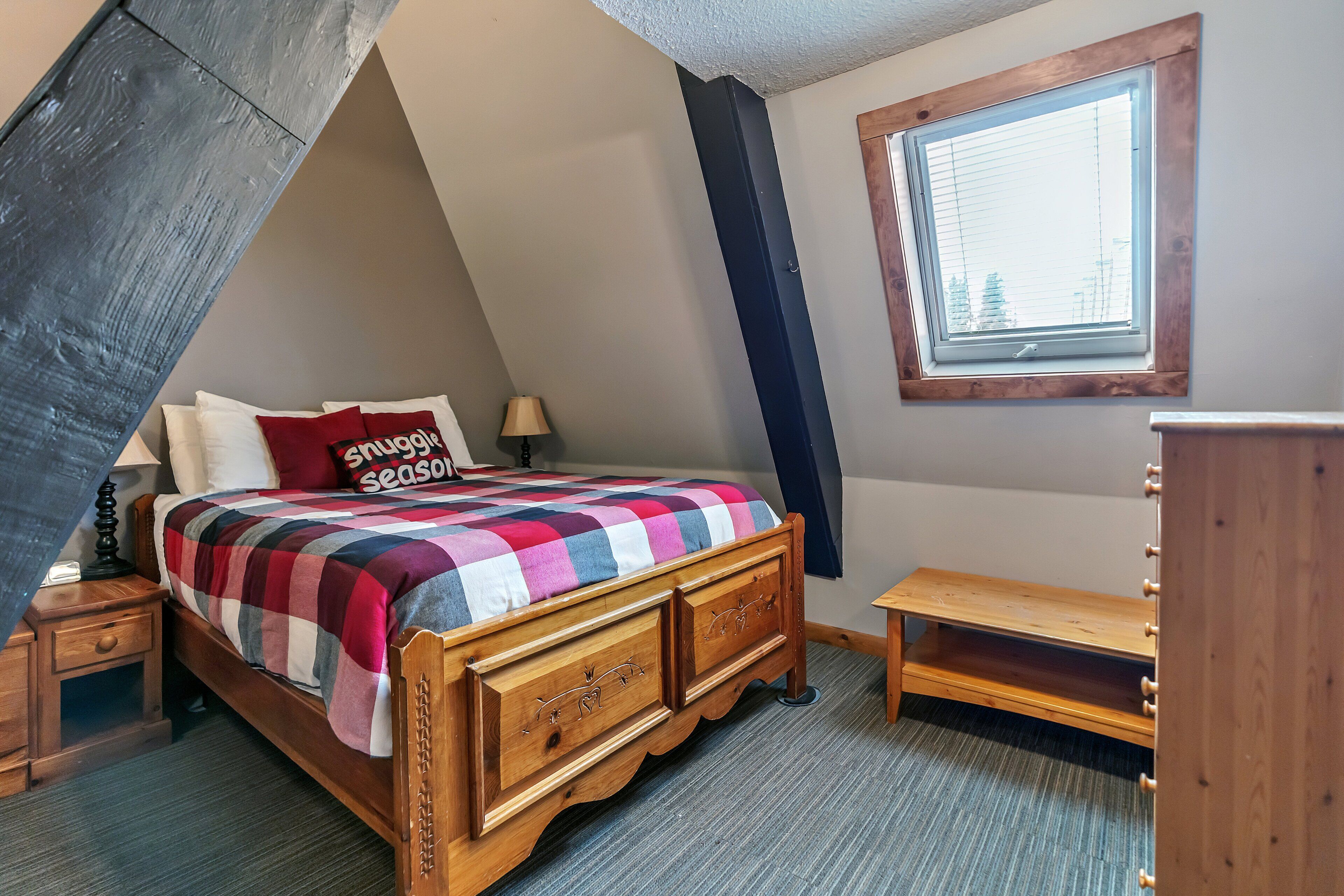 Une chambre avec un plafond en pente, un lit recouvert d’une couverture à carreaux, une table de chevet, une lampe, un banc en bois et une fenêtre avec des stores.